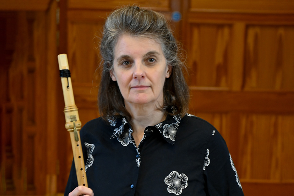 A woman wearing a patterned shirt, holding a historical clarinet, with a wooden panelled room.